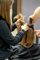 a traseiro Visão do dois cabeleireiro estão ondulação cabelo para uma jovem mulher com elétrico cabelo ferro dentro uma beleza salão. foto