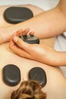 quente pedra massagem em a fêmea costas com mãos do massagista segurando Preto massagem pedras dentro spa salão. foto