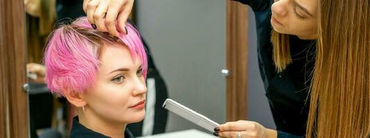 fêmea cabeleireiro estilo curto Rosa cabelo do a jovem branco mulher com mãos e pente dentro uma cabelo salão. foto