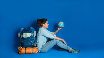 turismo de planejamento de férias com a ajuda do mapa-múndi com outros acessórios de viagem ao redor. viajante de mulher com mala sobre fundo de cor azul. garota com um globo de bola na mão. mochila de viagem foto