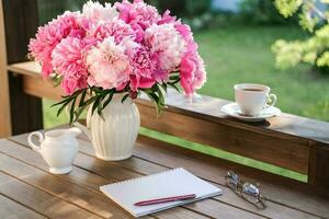 uma vaso com peônias, uma caderno e uma copo do café em a terraço dentro a verão jardim. foto