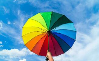 mão segurando colorida arco Iris guarda-chuva e azul céu fundo. lgbt, orgulho mês, diversidade, protetor solar conceito. foto