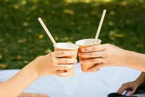dois crianças mãos estão segurando ecológico copos e tubos com maçã suco às uma piquenique dentro a parque. ecológico descartável talheres foto
