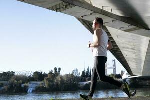 jovem homem corrida dentro uma branco camisa através uma ponte. ele é ouvindo para música e ele é obteve alguns capacetes sobre. foto