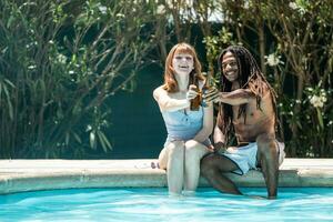 afro-americano homem e branco mulher brindar com Cerveja garrafas em a Beira do uma piscina. foto