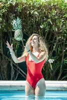 mulher dentro uma vermelho roupa de banho jogando uma abacaxi dentro a ar para dentro uma piscina foto