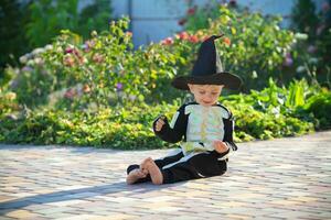 pequeno Garoto sentado em a Relva vestindo uma esqueleto traje e uma Preto chapéu. dia das Bruxas feriado. cópia de espaço foto