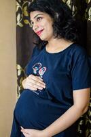 uma grávida indiano senhora poses para interior gravidez tiro e mãos em barriga, indiano grávida mulher coloca dela mão em dela estômago com uma maternidade vestir dentro dela casa foto