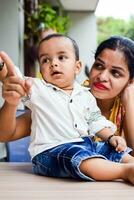 amoroso mãe carregando do dela bebê às casa sacada. brilhante retrato do feliz mãe segurando criança dentro dela mãos. mãe com dela pequeno 1 ano velho filho. foto