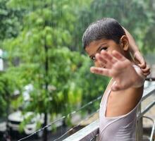 pequeno criança jogando dentro verão chuva dentro casa sacada, indiano inteligente Garoto jogando com chuva gotas durante monção chuvoso temporada, criança jogando dentro chuva foto