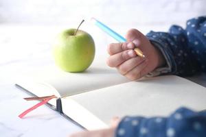 criança menina mão escrevendo no bloco de notas com maçã verde fresca na mesa foto