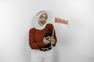 uma sorridente ásia muçulmano mulher vestindo vermelho topo e branco hijab, segurando dela telefone e bandeira, isolado de branco fundo. da indonésia independência dia conceito foto
