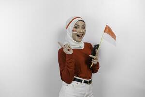 uma sorridente ásia muçulmano mulher vestindo vermelho topo e branco hijab, segurando dela telefone e bandeira, isolado de branco fundo. da indonésia independência dia conceito foto