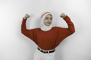 animado ásia muçulmano mulher vestindo uma vermelho topo e branco hijab mostrando Forte gesto de elevação dela braços e músculos sorridente orgulhosamente. da indonésia independência dia conceito. foto