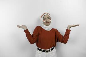 uma pensativo jovem muçulmano mulher vestindo branco hijab e vermelho topo é confundir entre escolhas ao lado dela, isolado de branco fundo. da indonésia independência dia. foto
