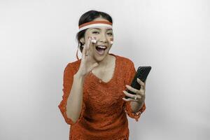 jovem lindo mulher vestindo uma vermelho kebaya é segurando dela telefone enquanto gritando e gritando alto com uma mão em dela boca. da indonésia independência dia conceito. foto