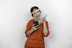 uma feliz ásia mulher vestindo vermelho kebaya e arco de cabelo, segurando dela telefone, e apontando cópia de espaço em topo do dela, isolado de branco fundo. da indonésia independência dia foto