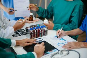 médico equipe tendo uma encontro com médicos dentro branco laboratório casacos e cirúrgico esfrega sentado às uma mesa discutindo uma pacientes trabalhando conectados usando computadores dentro a médico indústria foto