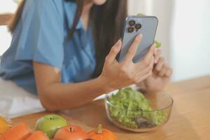 uma jovem mulher com uma lindo face dentro uma azul camisa com grandes cabelo comendo fruta sentado dentro a cozinha às casa com uma computador portátil e caderno para relaxamento, conceito período de férias. foto