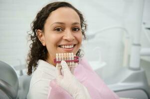 fechar-se do dentista mão segurando uma dentes cor gráfico perto a face do uma odontologia clínica mulher paciente sorridente com lindo cheio de dentes sorrir olhando às Câmera. dentes branqueamento, branqueamento conceito foto