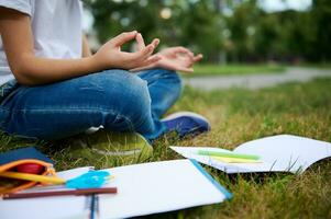 cortada Visão do escola criança Garoto sentado dentro lótus posição em verde Relva do cidade parque e meditando . livros de exercícios escola suprimentos deitado em a grama. concentração, lazer, atenção conceitos foto