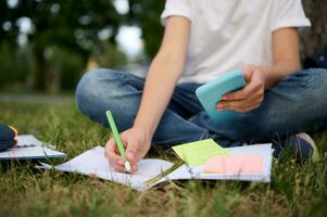 cortada imagem do a irreconhecível escola Garoto estudando dentro a parque, sentado em verde Relva e resolução matemática tarefa, usando Smartphone e Móvel formulários, fazer notas em caderno e pasta de trabalho. foto