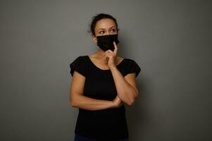 isolado retrato em cinzento fundo do uma mulher dentro Preto médico mascarar com cruzado braços olhando pensativamente para a lado às uma cópia de espaço para anúncio. Preto Sexta-feira conceito foto