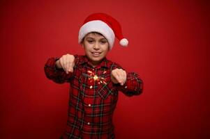 bonito adorável pré-adolescente garoto, bonita criança dentro santa claus chapéu e xadrez camisa goza a Natal festa, tocam com Bengala luzes, estrelinhas, isolado sobre vermelho fundo com cópia de espaço para de Anúncios foto