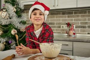 adorável bonito pré-adolescente Garoto dentro xadrez camisa e santa chapéu detém uma de madeira colher e carrinhos de cozinha mesa com uma tigela com Natal biscoitos massa, sorrisos cheio de dentes sorrir olhando às Câmera foto