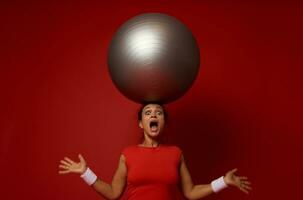 atônito jovem desportivo misturado raça mulher poses contra vermelho parede fundo com enorme ginástica bola em dela cabeça foto