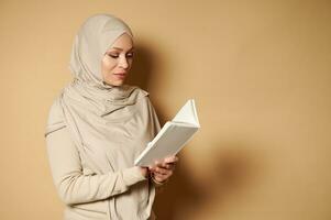 muçulmano árabe mulher vestindo hijab e rigoroso formal tradicional equipamento lendo uma livro em bege fundo com cópia de espaço foto