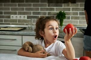 lindo bebê menina abraçando dela suave pelúcia brinquedo e olhando às maduro vermelho tomate dentro dela mão sentado às uma cozinha mesa foto