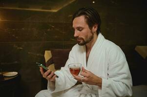relaxado bonito atraente meio envelhecido caucasiano homem dentro roupão de banho bebendo chá e olhando às uma Smartphone dentro dele mão, navegando, lendo notícia enquanto em repouso de jacuzzi às bem estar spa recorrer foto