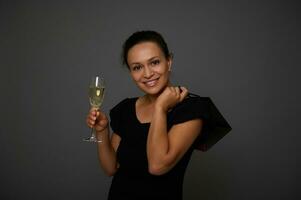 lindo mulher comprador vestido dentro Preto vestir fofinho sorrisos olhando às Câmera posando contra cinzento fundo com uma vidro do vinho e compras Preto pacote. conceito do celebração a Preto Sexta-feira foto