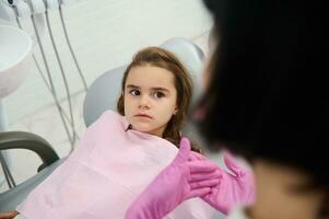 pequeno menina sentado dentro dentista cadeira, olhando às uma pediatra higienista ensino dela sobre oral higiene e prevenção do bebê dentes decair durante dental Visita dentro branco limpar \ limpo moderno médico gabinete foto