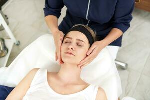 jovem atraente Loiras mulher desfrutando facial massagem durante Visita para cosmetologista gabinete foto