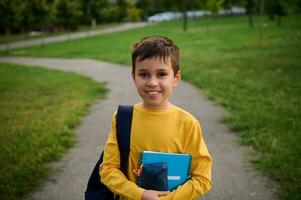 uma bonito estudante com uma escola saco e cadernos carrinhos em a caminho dentro a cidade parque depois de escola. retrato do uma fofa adorável estudante foto