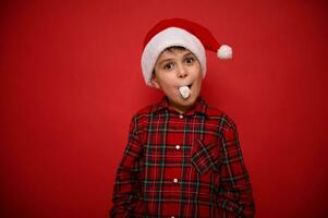 lindo surpreendente engraçado e alegre pré-adolescente garoto, vestindo santa chapéu e xadrez vermelho camisa parece às Câmera posando contra colori fundo com açucarado marshmallow dentro dele boca. Natal conceito foto