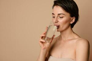 atraente caucasiano nu natural beleza mulher com saudável brilhando limpar \ limpo pele bebendo água, isolado em bege fundo com cópia de espaço para Publicidades. hidratação, pele e corpo Cuidado, refresco conceito foto