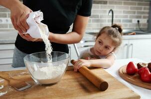 mulher derramando branco farinha a partir de papel embalagem para dentro uma transparente vidro tigela. Próximo para dela senta dela pequeno filha e relógios a cozinhando processo. foto