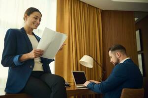 lindo meio envelhecido bem sucedido Deleitado mulher dentro casual terno sentado em uma mesa com documentos enquanto dela parceiro de negócios, digitando em computador portátil teclado trabalhando juntos dentro hotel quarto foto