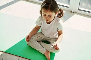 adorável menina com rabos de cavalo é sentado em a exercício esteira e fazendo meditação. em desenvolvimento saudável hábitos a partir de cedo infância. foto