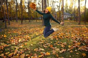 lindo mulher dentro caloroso verde estacionar, amarelo de lã e casual jeans chapéu pulando Alto com uma ramalhete do lindo seco outono bordo folhas dentro mãos, desfrutando lindo outono dia às pôr do sol dentro parque foto