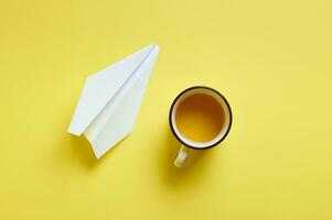 plano deitar composição do uma branco copo do café ou cappuccino e uma papel avião isolado em uma amarelo fundo. topo visualizar, cópia de espaço foto