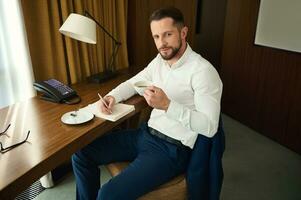 bonito autoconfiante homem de negocios, empreendedor em o negócio viagem bebendo café e escrevendo em bloco de anotações, sentado às uma escrivaninha dentro hotel quarto foto