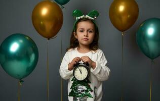 adorável pequeno menina vestindo a duende aro detém a alarme relógio com meia noite em a discar, parece às Câmera posando sobre cinzento fundo decorado com dourado verde ar balões. alegre Natal conceito foto