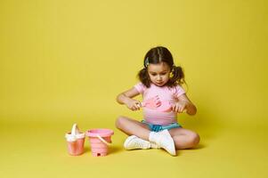 adorável pequeno menina jogando com pá e ancinho, sentado perto uma conjunto do de praia brinquedos. isolado verão conceitos em amarelo fundo foto