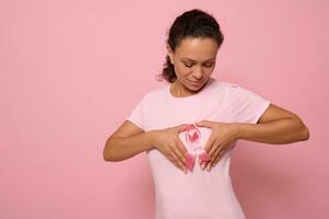 isolado retrato em colori fundo do africano americano mulher dentro Rosa camiseta, colocando dela mãos em dela peito dentro forma do coração com uma Rosa cetim fita dentro a Centro. mundo Câncer consciência dia. foto