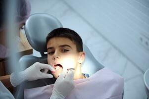 escola Garoto às dental encontro. a sobrecarga Visão do pediatra dentista examinando oral cavidade do uma Adolescência Garoto sentado em dental cadeira, examina a dentes do a presença do cárie usando uma dental espelho. foto