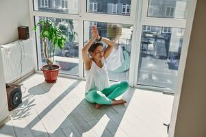 atento jovem mulher dentro roupa de esporte sentado dentro padmasana lótus posição com elevado braços desfrutando fechadas olhos meditação durante ioga prática às casa foto